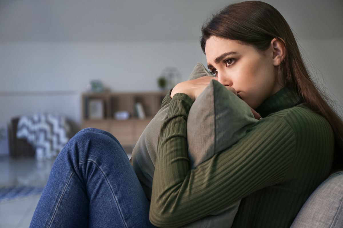Sad young white woman at home sitting on a couch with knees to chests holding a pillow for comfort