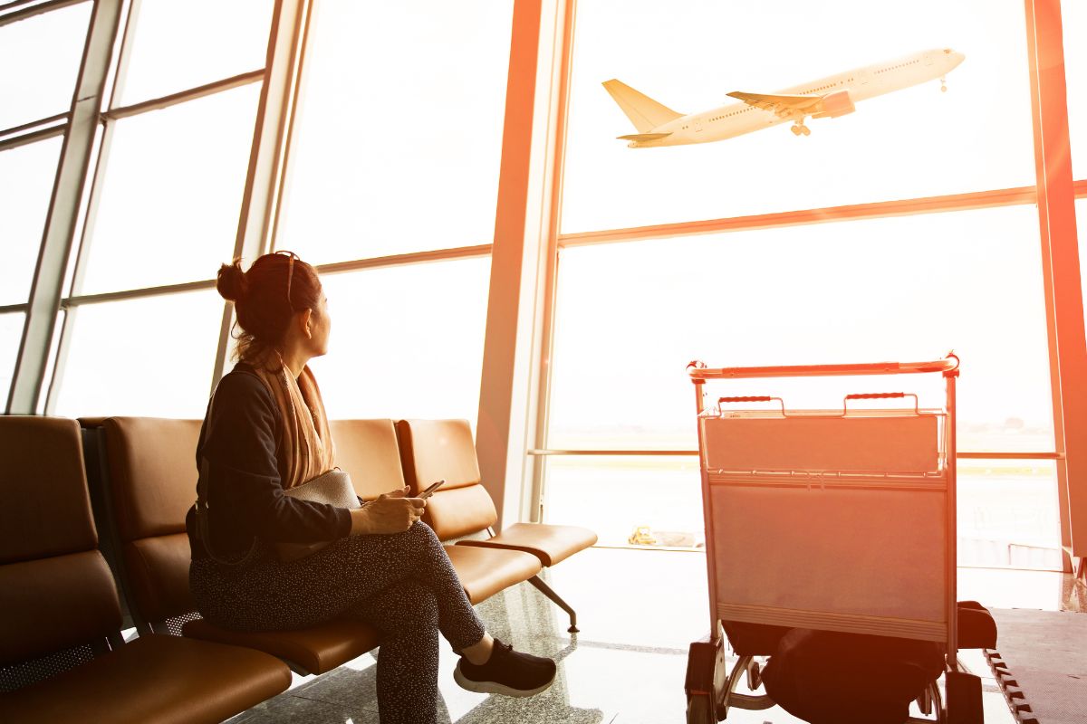 Single woman sitting in airport terminal looking out the window at a passenger plane taking off outside.