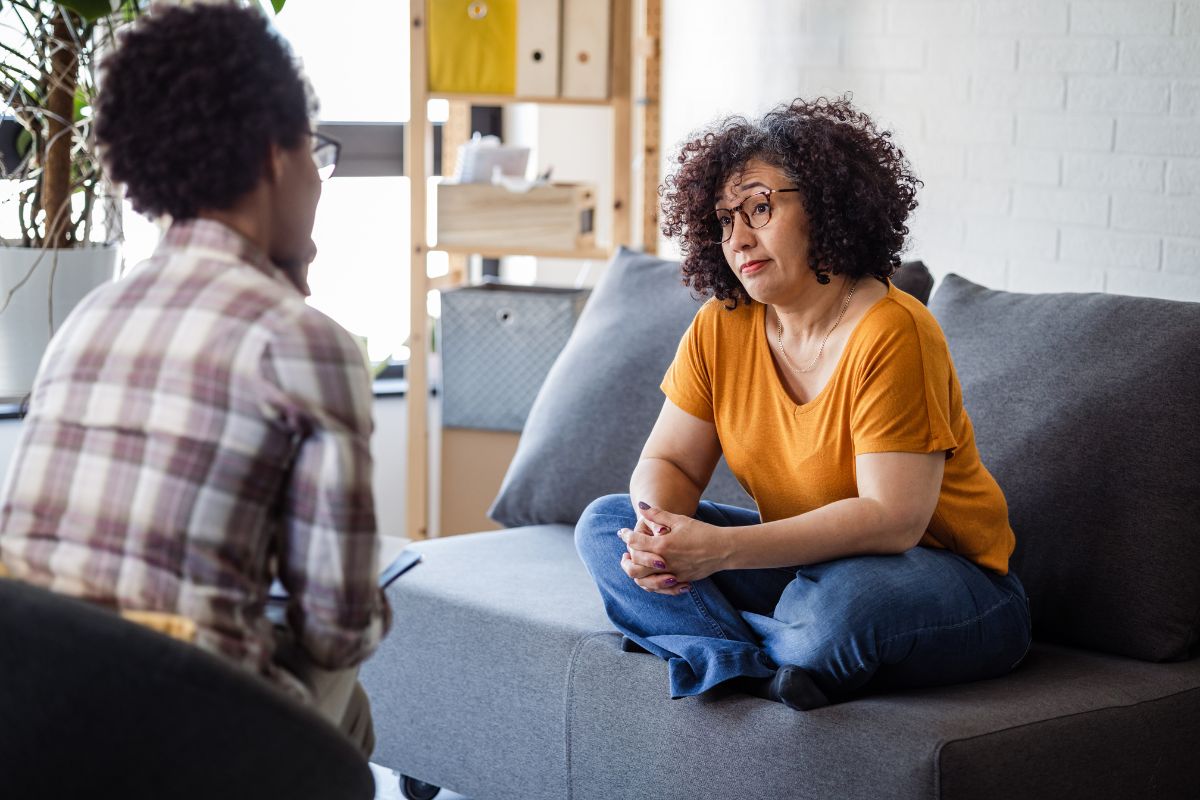 A therapist with back to viewer sitting across from a client who is sitting crossed legged and hands clasped over legs on a couch looking quisitively.