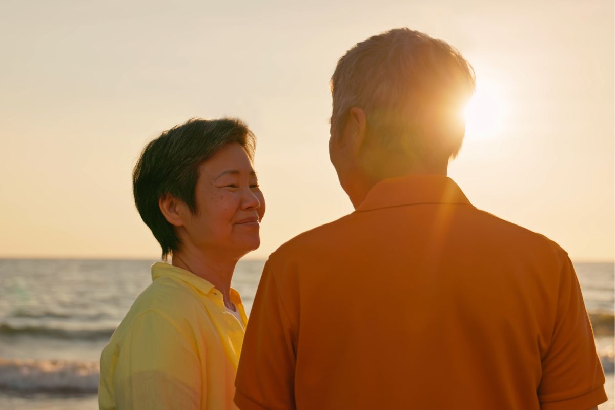 Older Asian couple standing on a beach at sunset looking at each other and smiling.