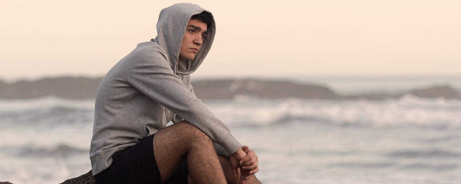 Worried hispanic teenager sitting on rocks beside the sea. 