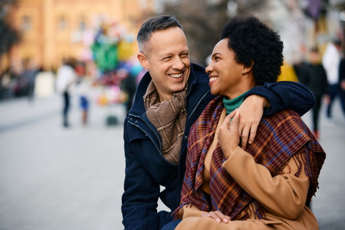 Interracial couple walking through the city on a wintery day with the man's arm around the woman shoulder smiling at each other and walking