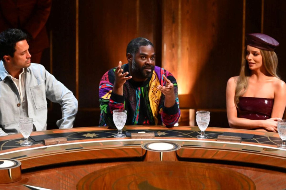 Ron Funches sitting at the round table talking with his hands while cast members on either side look at him.
