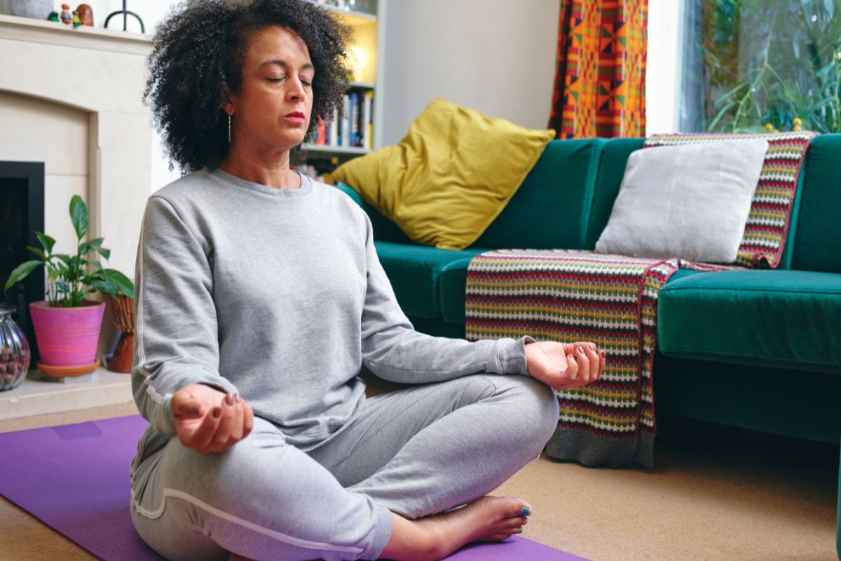 Middle aged black woman sitting on purple yoga mat in living room mediating
