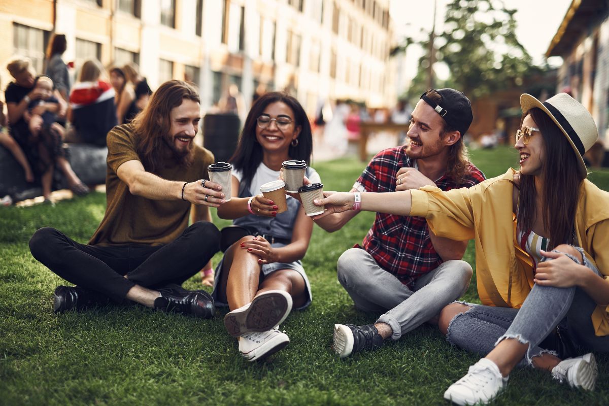  Positive young friends wearing casual clothes and sitting on the grass with cups of coffee in their hands and smiling