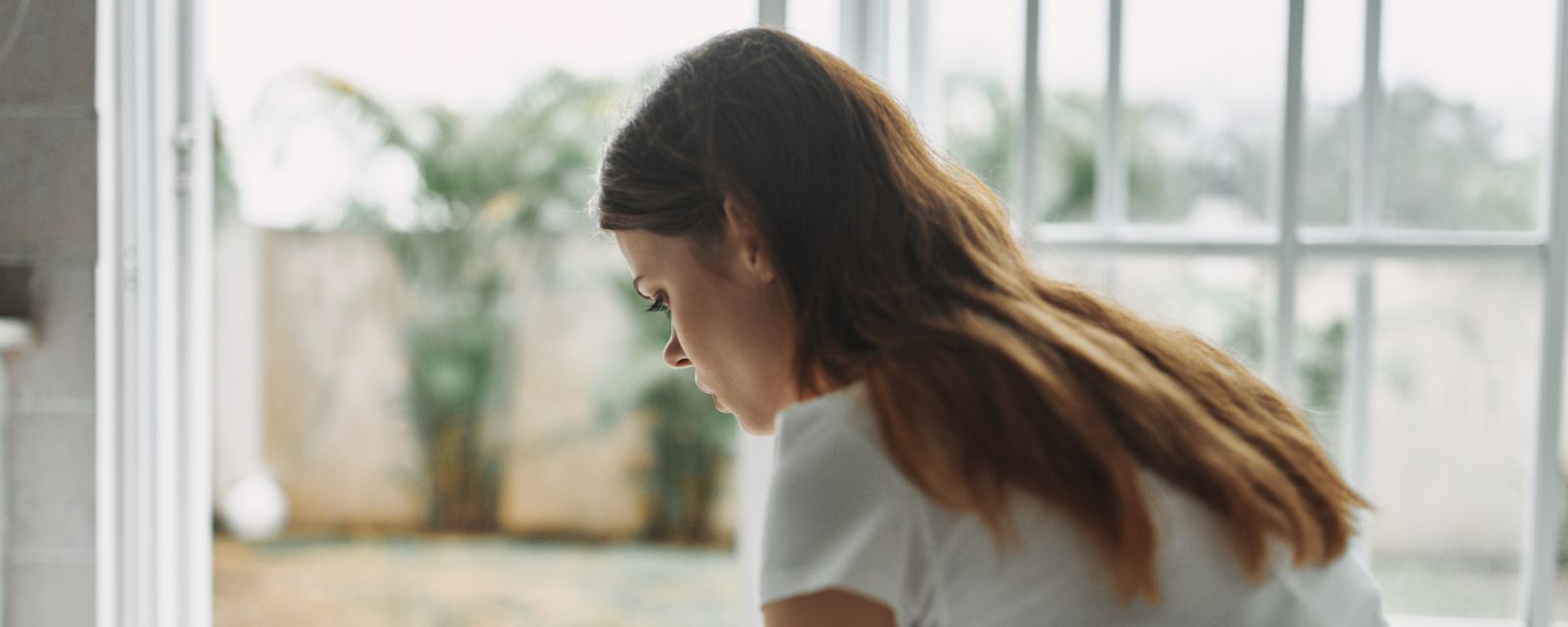 Young White woman sitting on a couch inside looking downward and sad with a door open to the outside that is warm and sunny