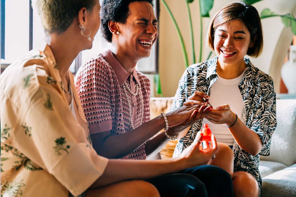 Three diverse friends sitting on a couch laughing, having fun while the person in the middle is getting their nails painted by the friend on the right while the friend on the left holds the nail polish bottle.
