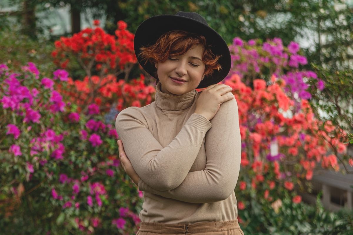 A beautiful mid size woman in a hat hug herself enjoying standing among the green plants of the greenhouse.