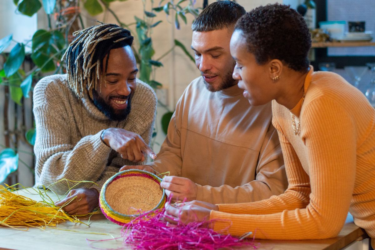 Ethically diverse group of three friends gathering around a table weaving baskets talking with and smiling at one another.