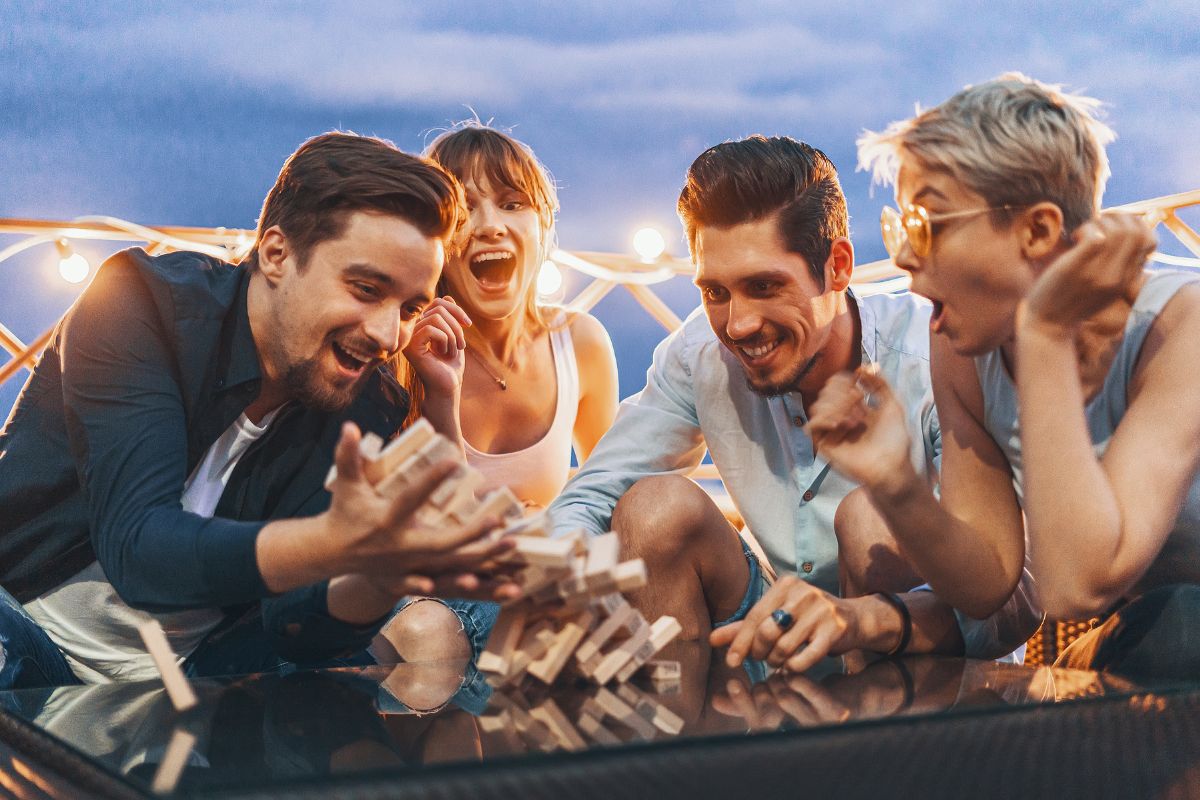 Group of young people playing jenga on patio during the early evening. Jenga tower is falling over and group is laughing, excited, shocked and all are having fun.