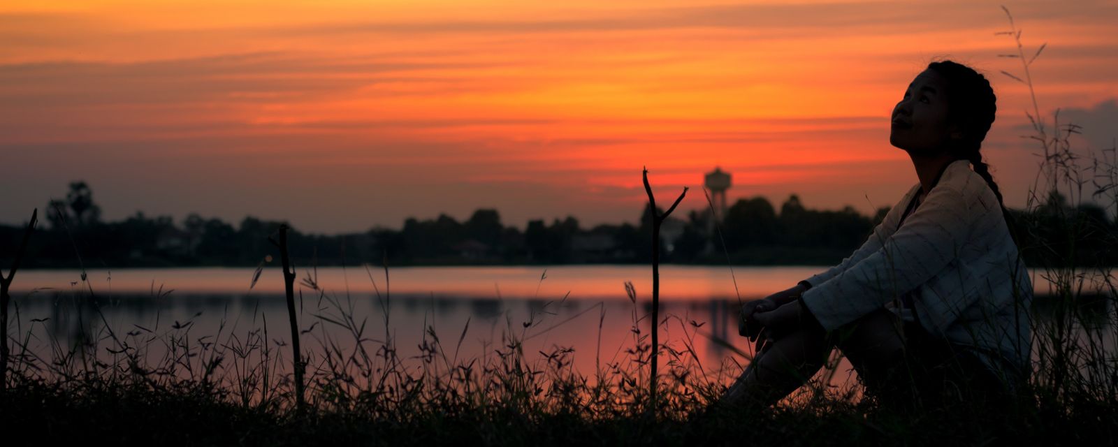 Silhouette of woman sitting and looking sky during an orange sunset over a pond