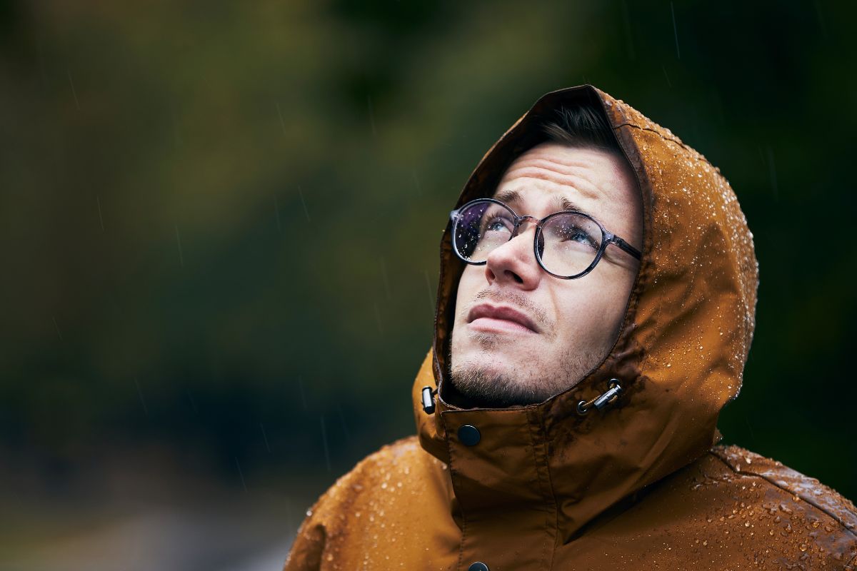 White man in waterproof jacket looking up at the sky with a worried face while it rains