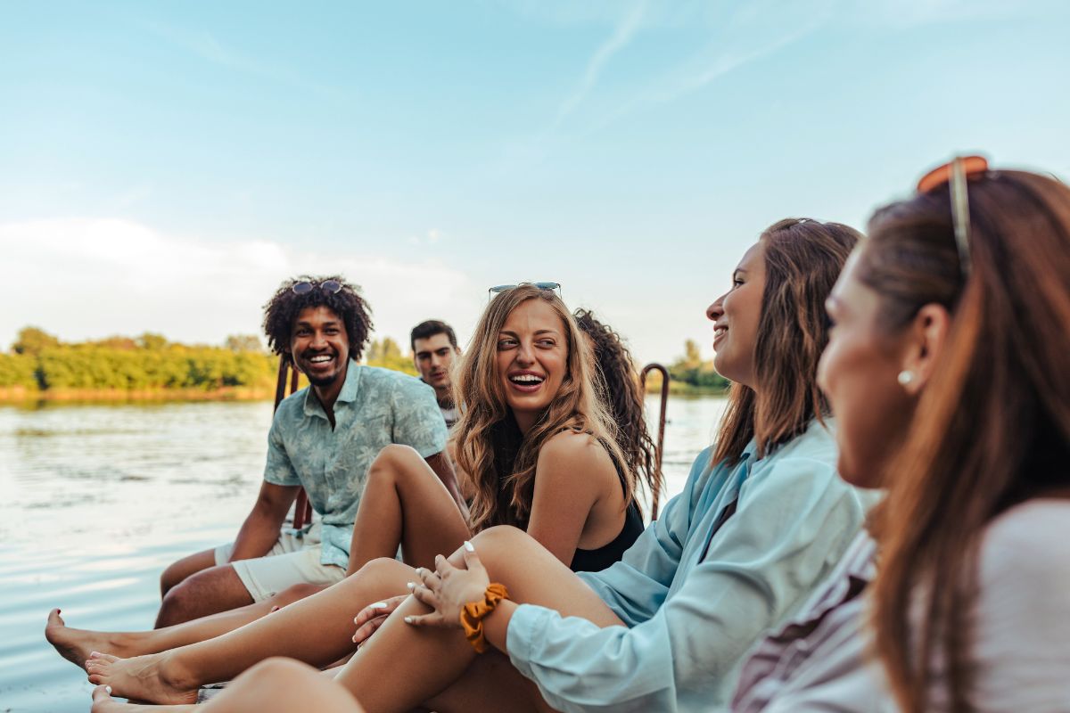 A group of diverse friends sits together outdoors near a calm lake at golden hour, laughing and talking.