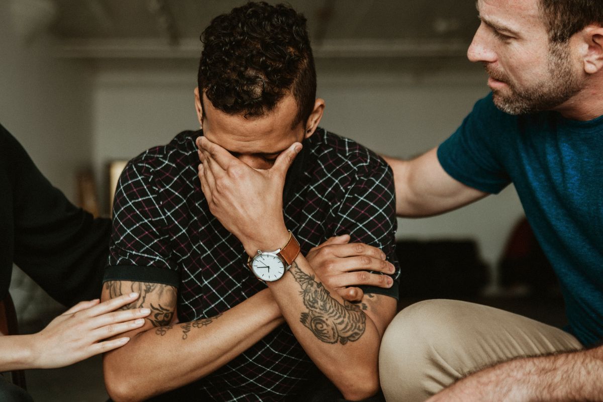 Man sad with his hand cover his face while man to the right along with a woman's hand to the left is comforting him 