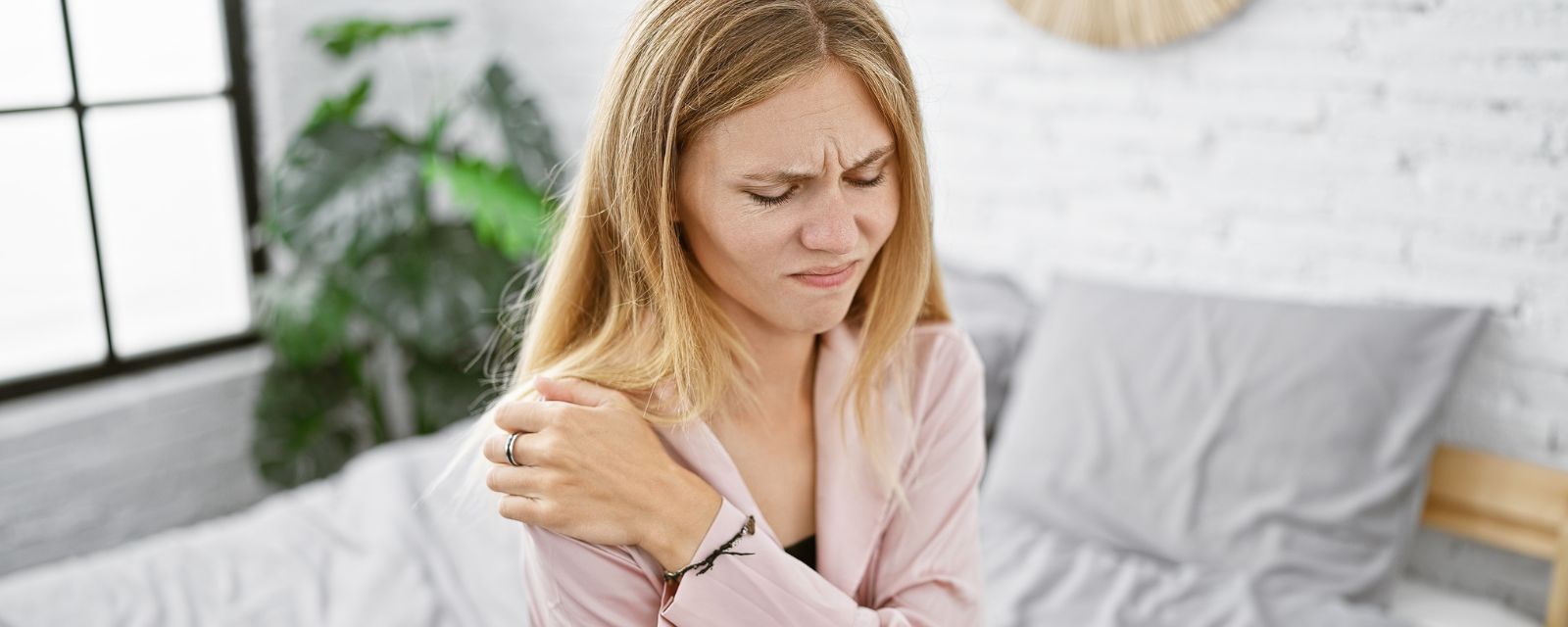 Blonde woman expressing pain while holding her shoulder in a bright bedroom setting