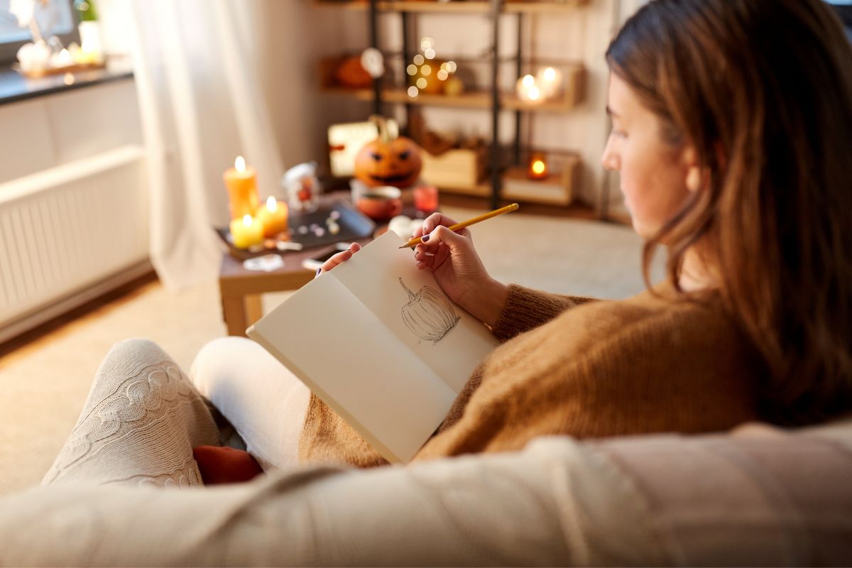 Young woman with pencil drawing pumpkin in sketchbook or diary at cozy home