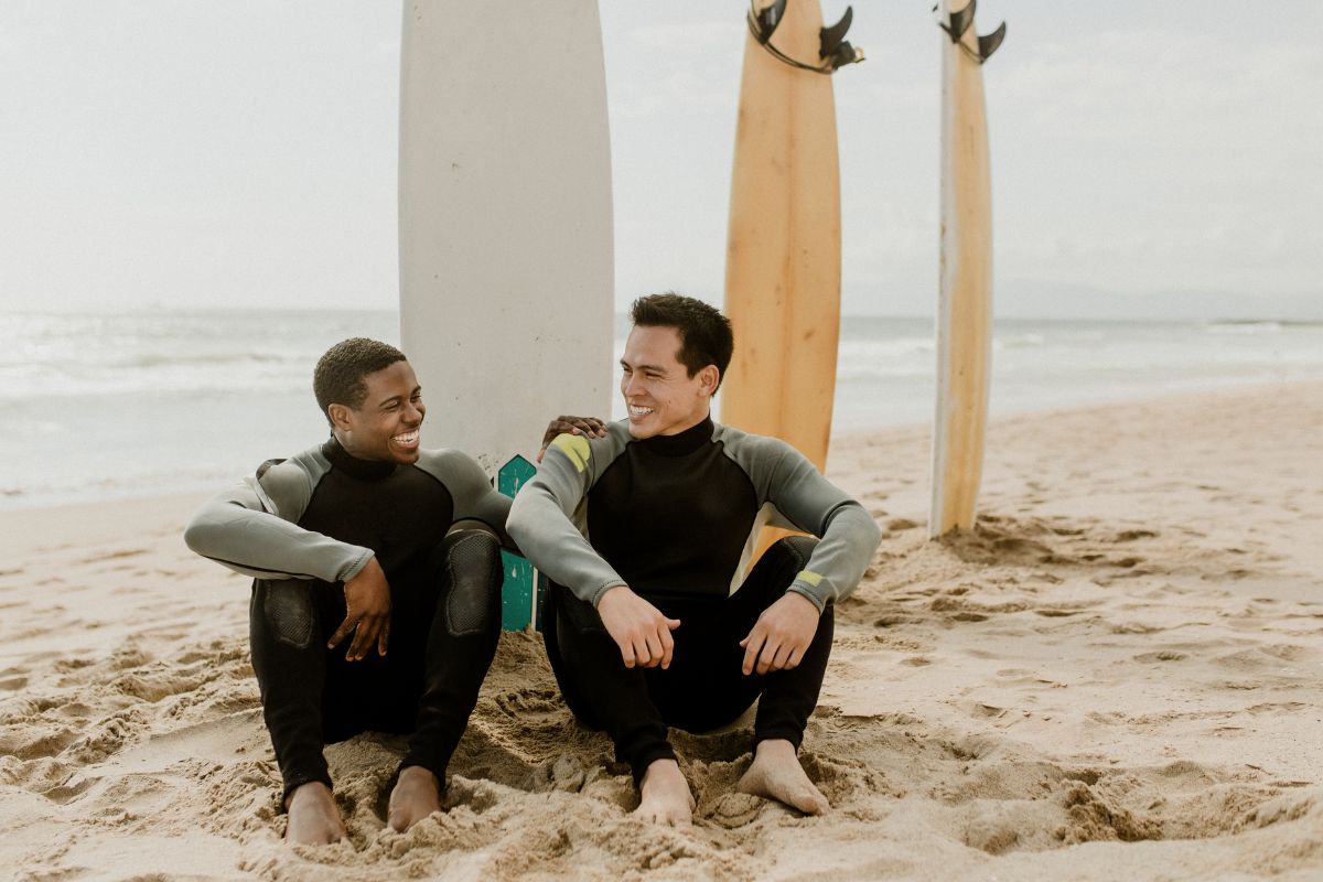 Two surfer friends sitting on the sand at the beach with their backs to the waves and their surfboards standing upright in the sand 