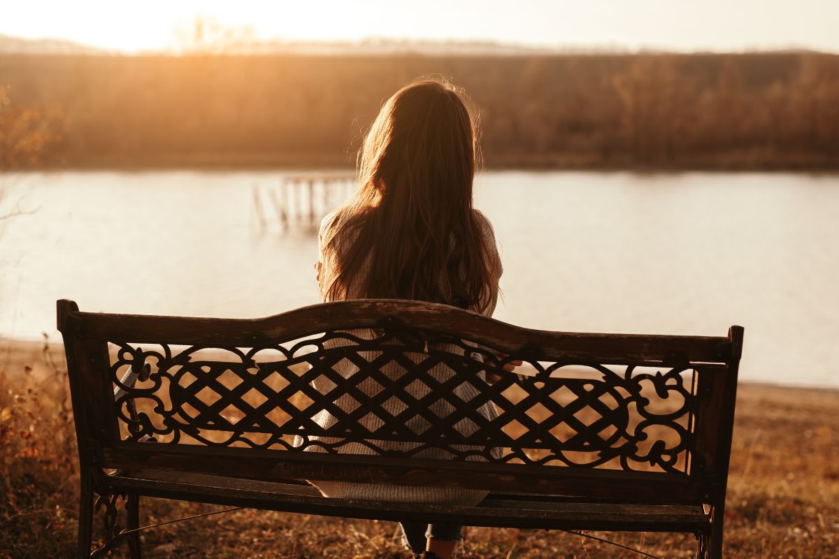 Young woman sitting alone on park bench by a river at sunset 