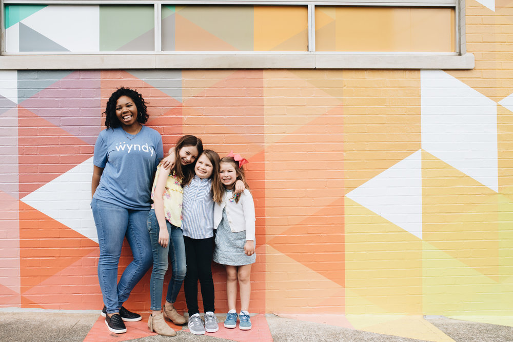 A sitter in a Wyndy t-shirt standing with three children in front of a colorful mural
