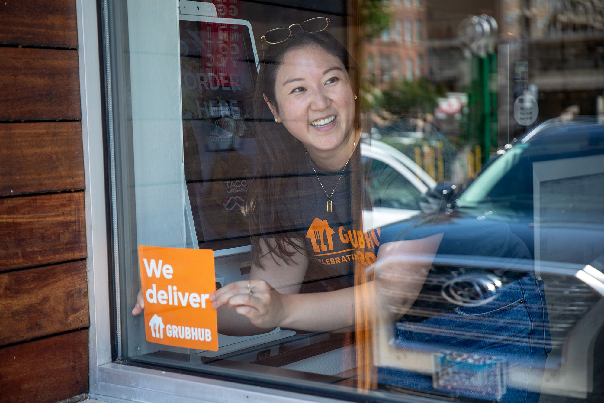 A restaurant worker in a Grubhub t-shirt holding a We deliver sign in the window, next to an ordering tablet