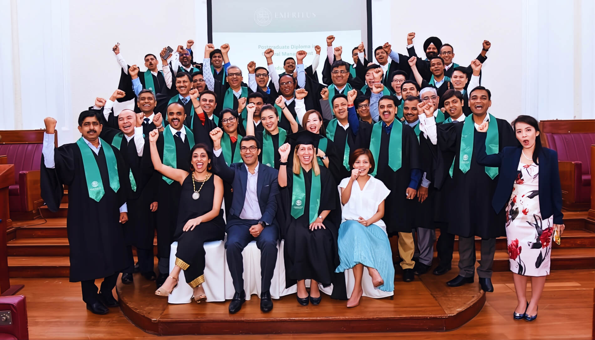 Emeritus graduates in gowns and green stoles cheering on stage at a postgraduate diploma ceremony