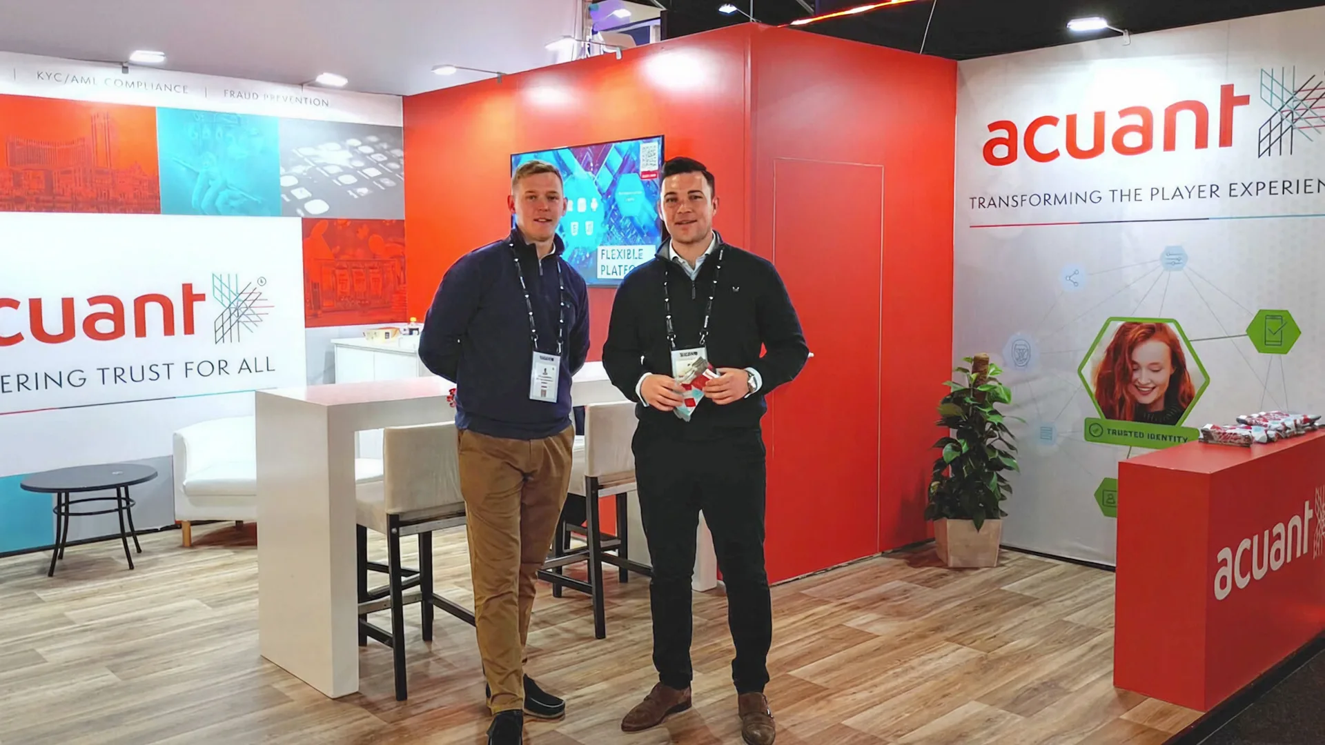 Acuant's trade show booth with signage for KYC/AML compliance and fraud prevention, and two people standing in front of it.