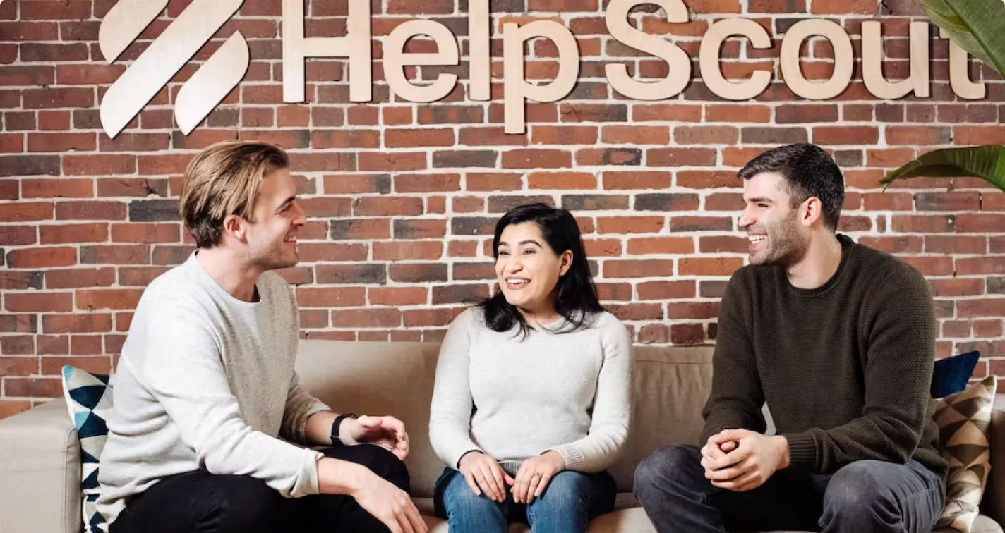 Three Help Scout employees talking on a couch in front of a wooden Help Scout logo on a brick wall