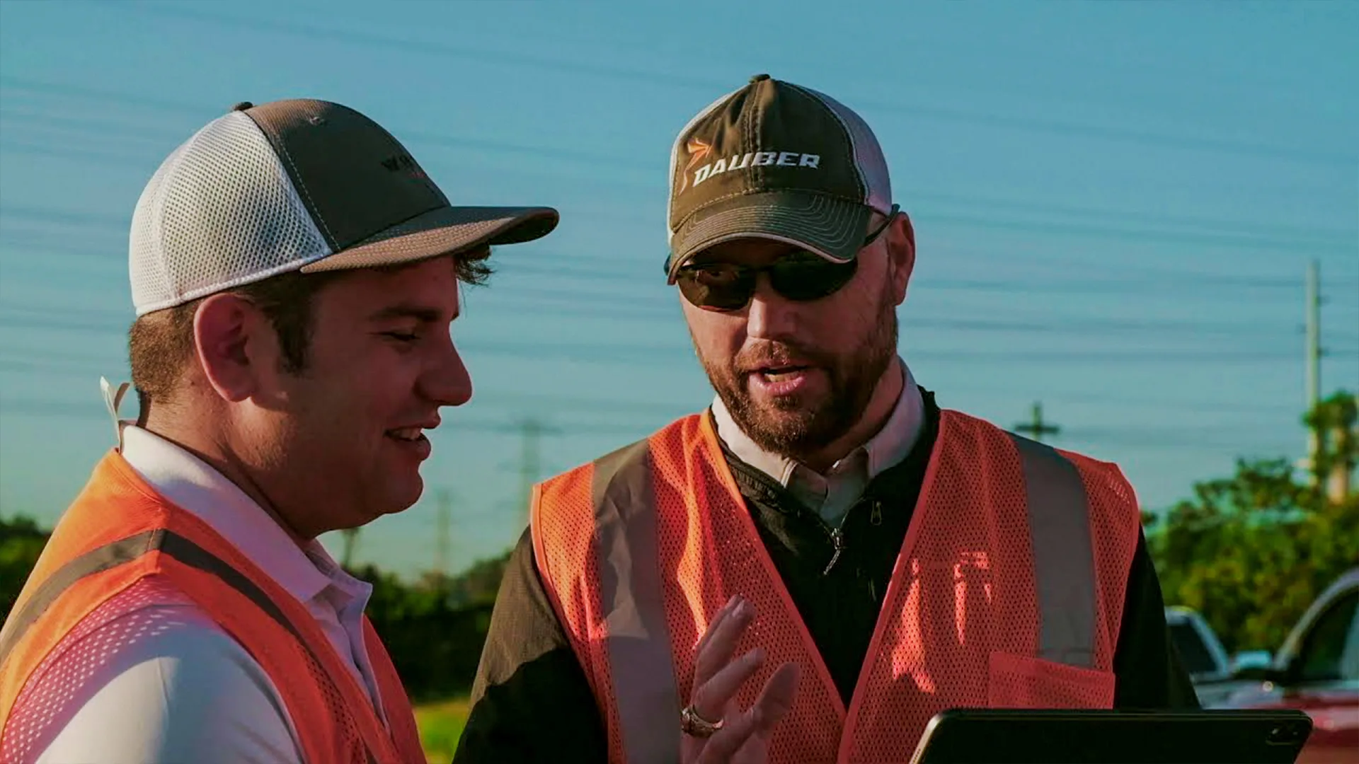 Two workers in high-visibility vests on a construction site looking at the Dauber app on a tablet, one wearing a Dauber cap