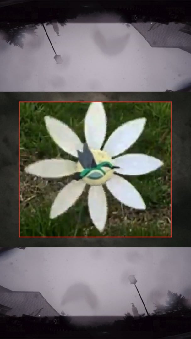 A white daisy-shaped pinwheel with a bird in the center set against grass, framed by a stormy porch view.