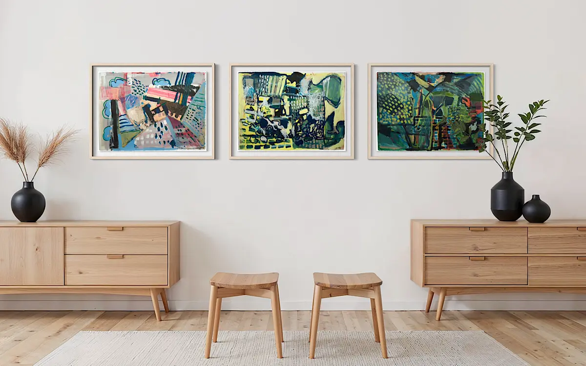 Three framed abstract paintings hang side by side above a light wood credenza in a modern minimalist living room. The artworks feature layered shapes, patterns, and brushstrokes in blue, green, yellow, and pink tones. Two simple wooden stools sit below them, with black vases holding dried and leafy plants on each end of the cabinet, creating a balanced, airy composition.
