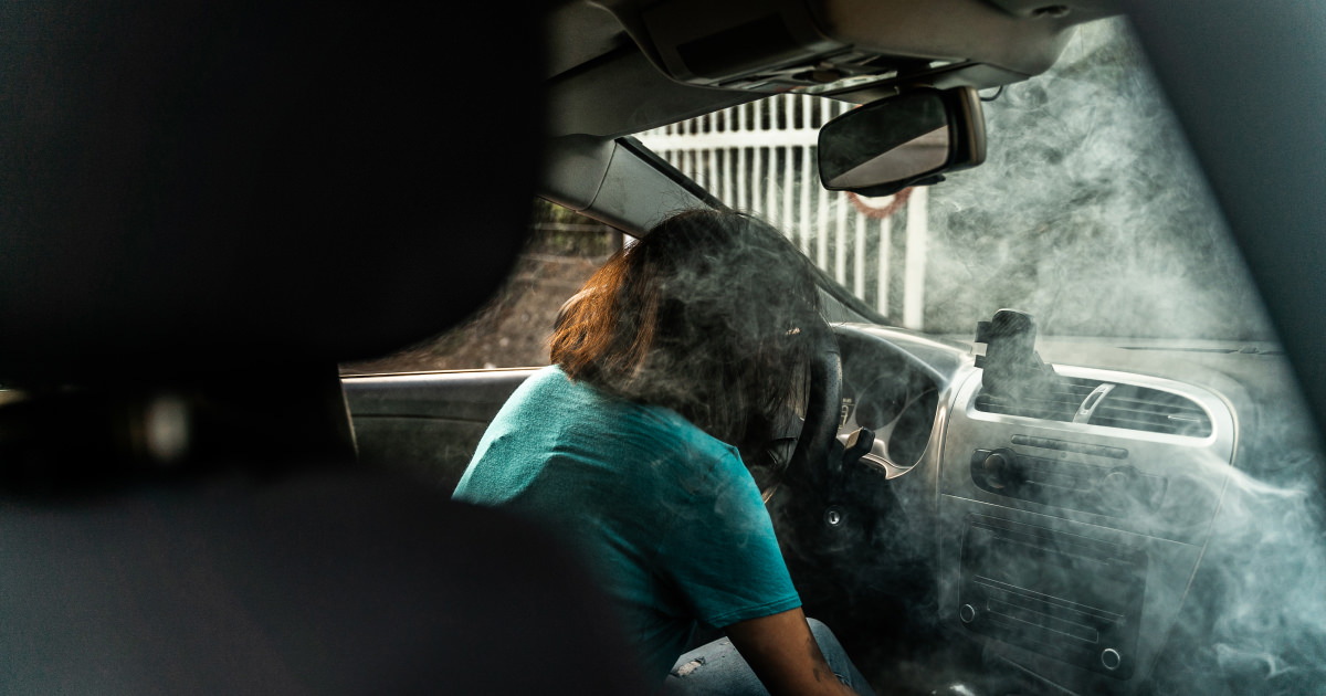 Person with brown hair slumped over the steering wheel inside a smoke-filled car