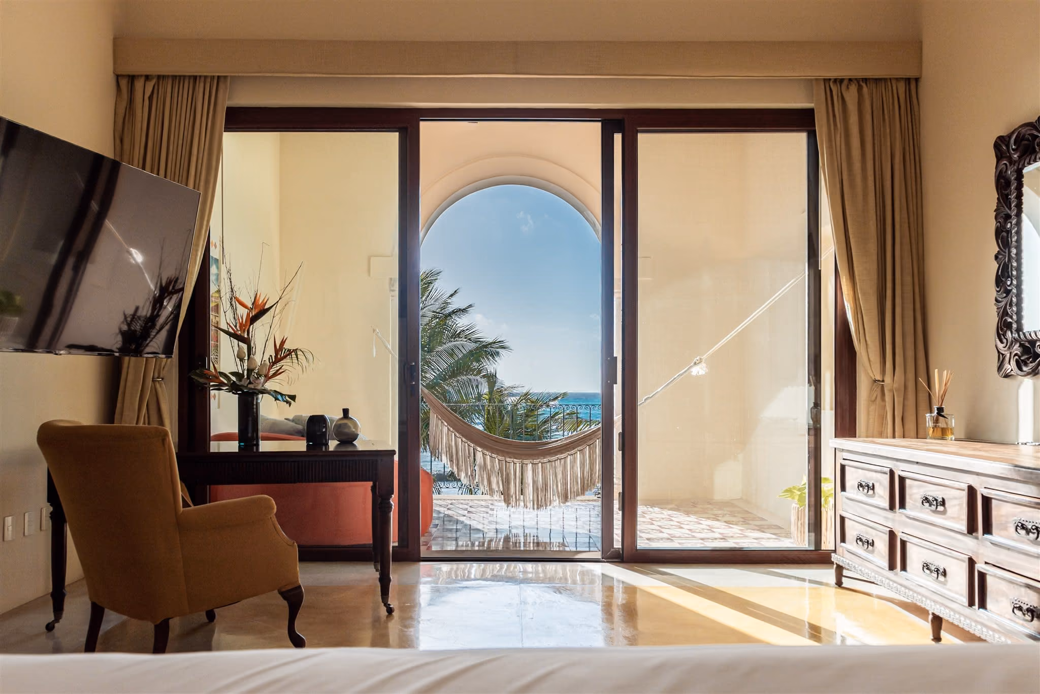 Outdoor pool with clear blue water, surrounded by palm trees, beige cushioned seating, and ocean view in the background.
