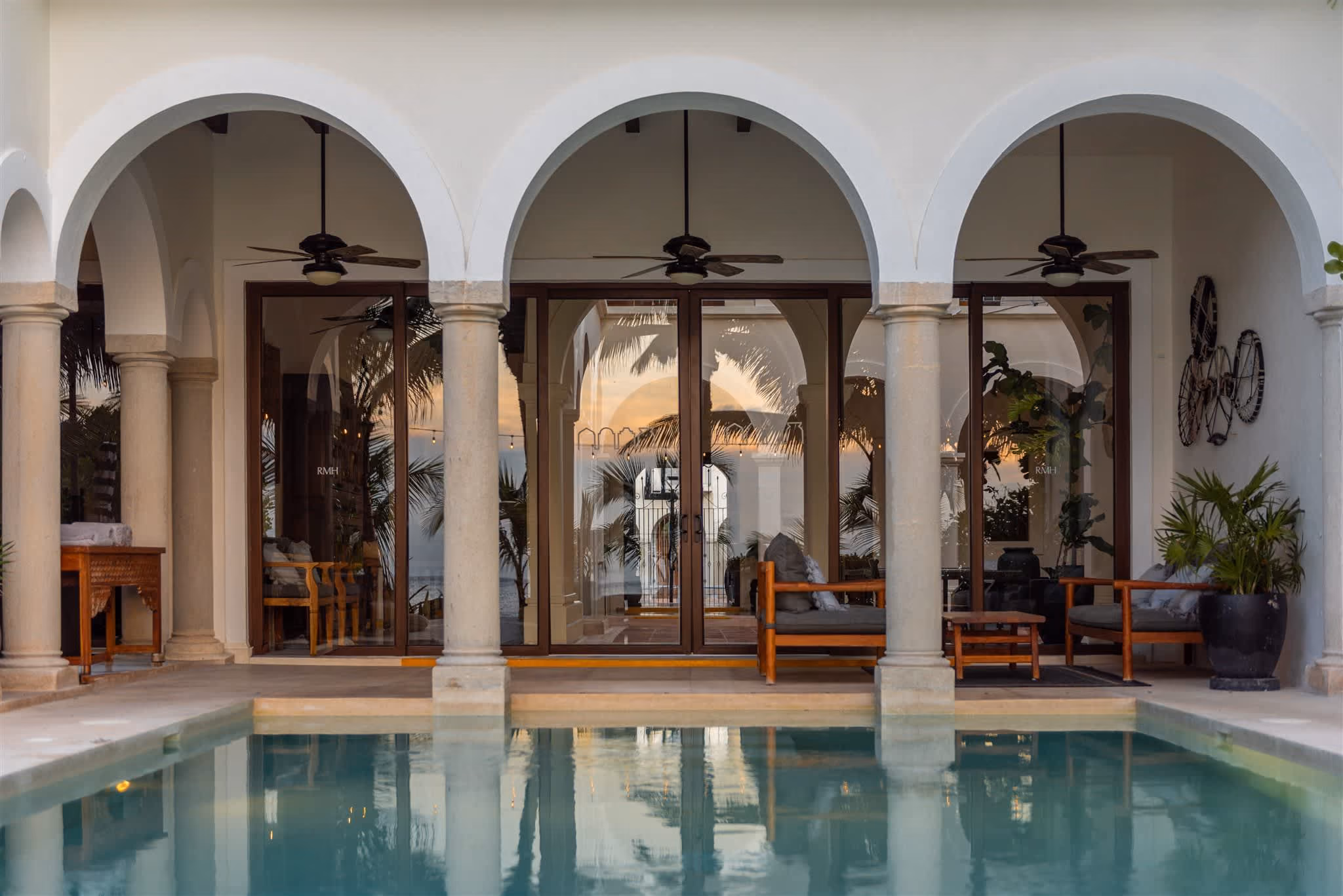 Outdoor pool with clear blue water, surrounded by palm trees, beige cushioned seating, and ocean view in the background.