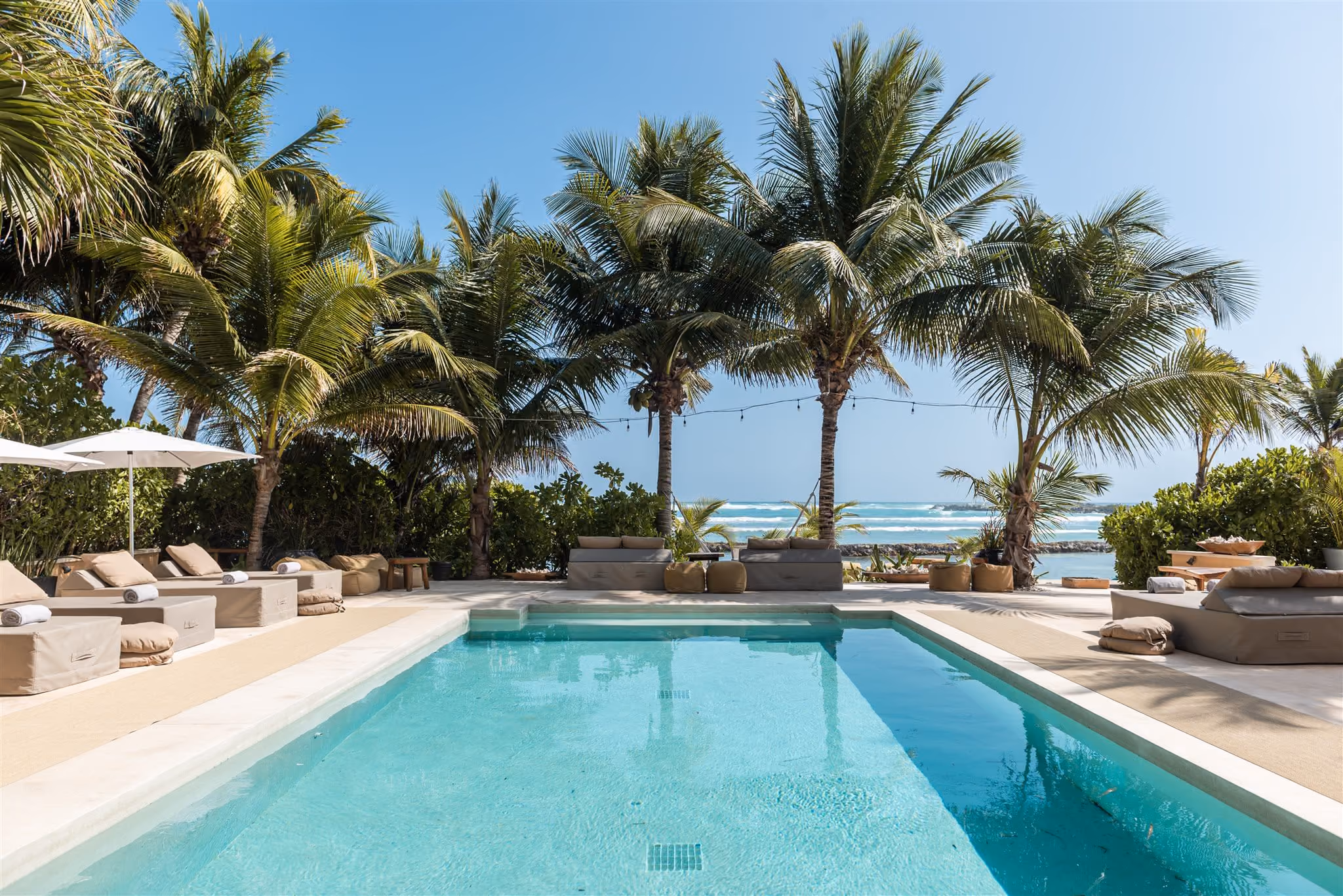 Outdoor pool with clear blue water, surrounded by palm trees, beige cushioned seating, and ocean view in the background.