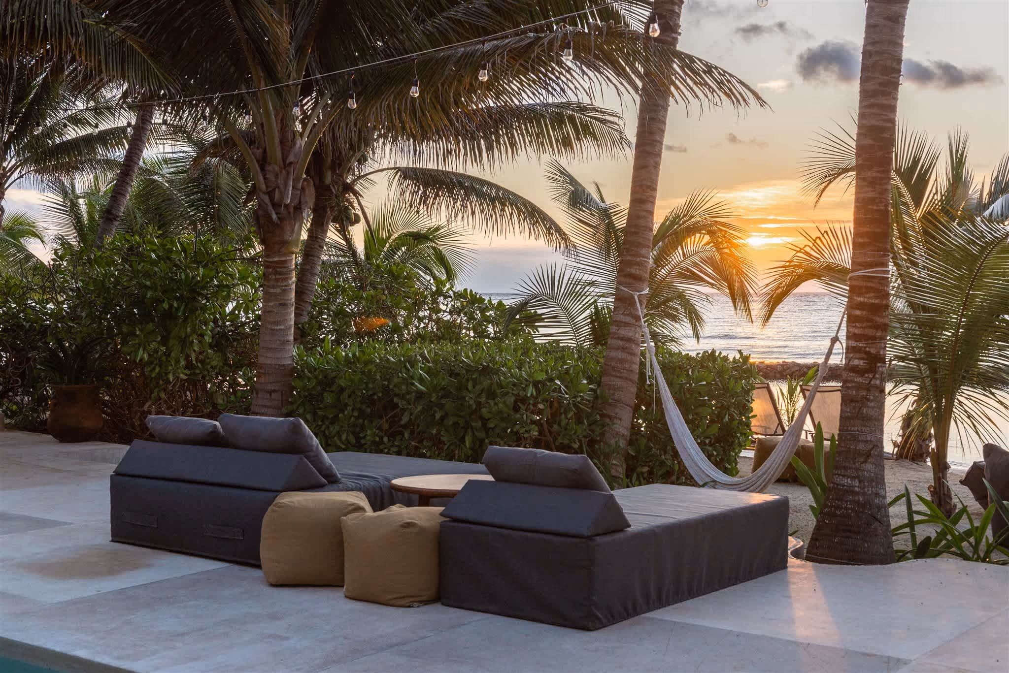 Outdoor pool with clear blue water, surrounded by palm trees, beige cushioned seating, and ocean view in the background.