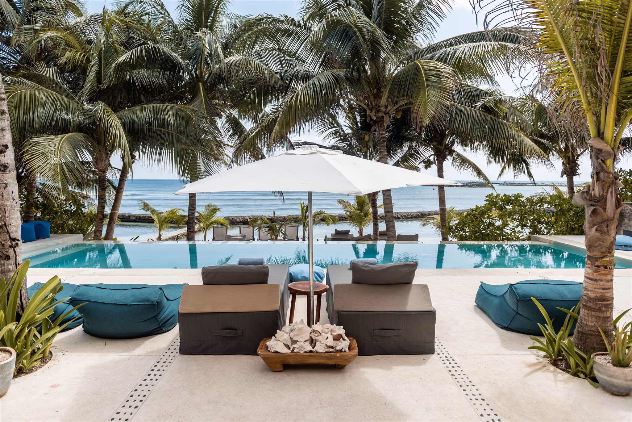 Outdoor pool with clear blue water, surrounded by palm trees, beige cushioned seating, and ocean view in the background.