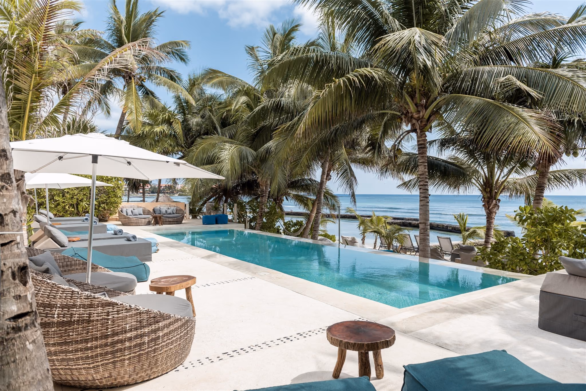Outdoor pool with clear blue water, surrounded by palm trees, beige cushioned seating, and ocean view in the background.