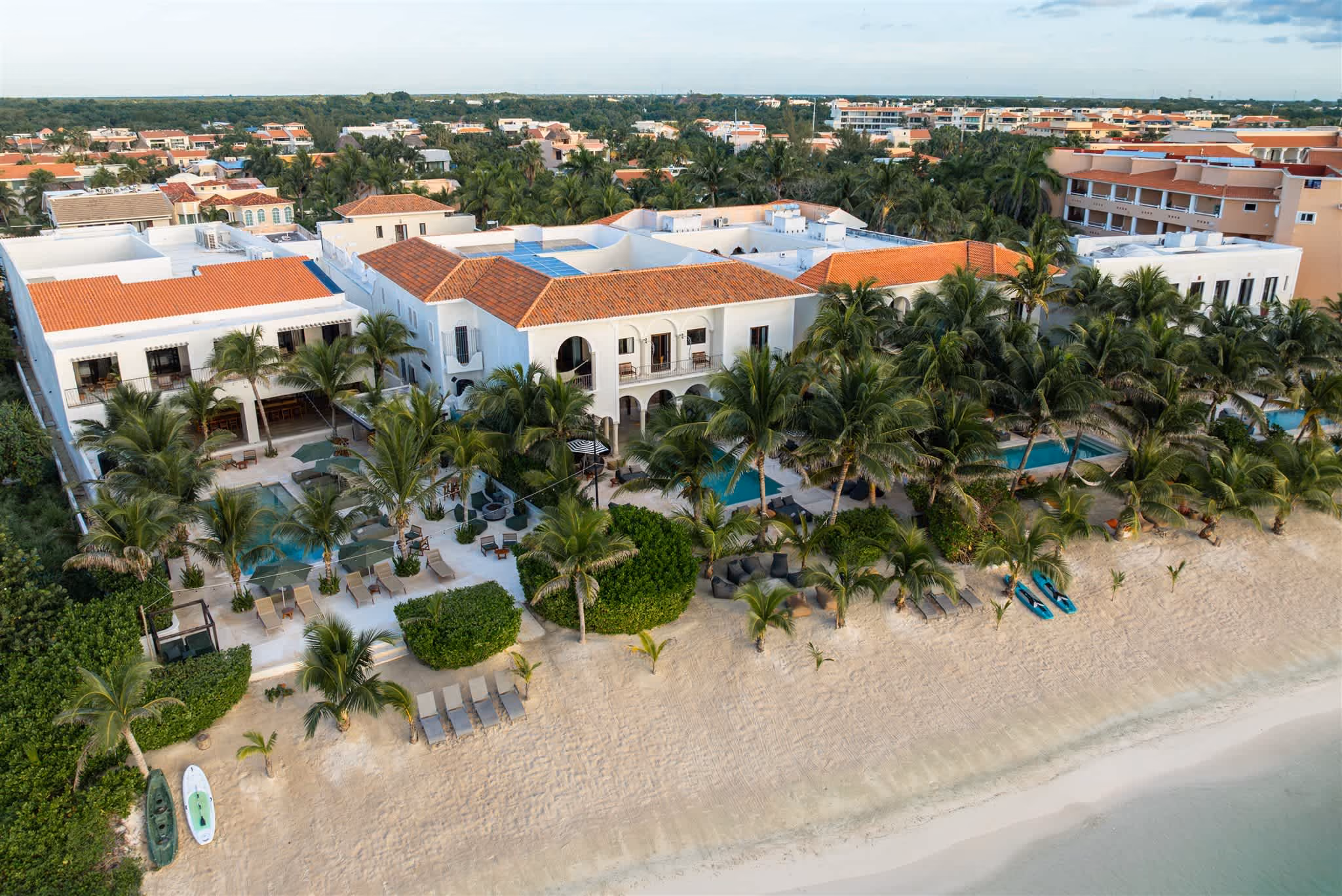 Outdoor pool with clear blue water, surrounded by palm trees, beige cushioned seating, and ocean view in the background.
