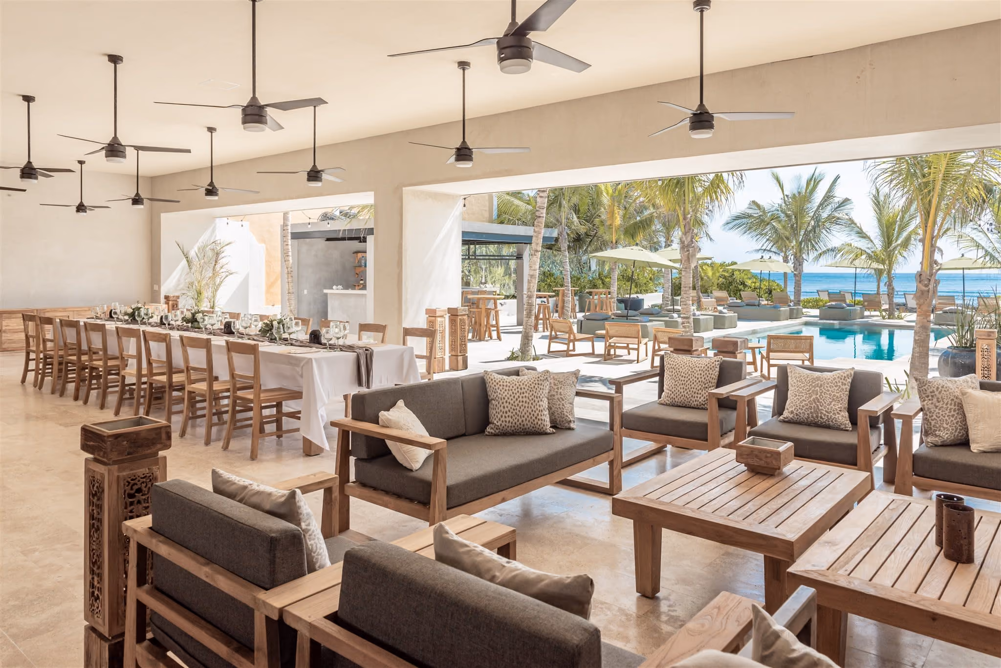 Outdoor pool with clear blue water, surrounded by palm trees, beige cushioned seating, and ocean view in the background.