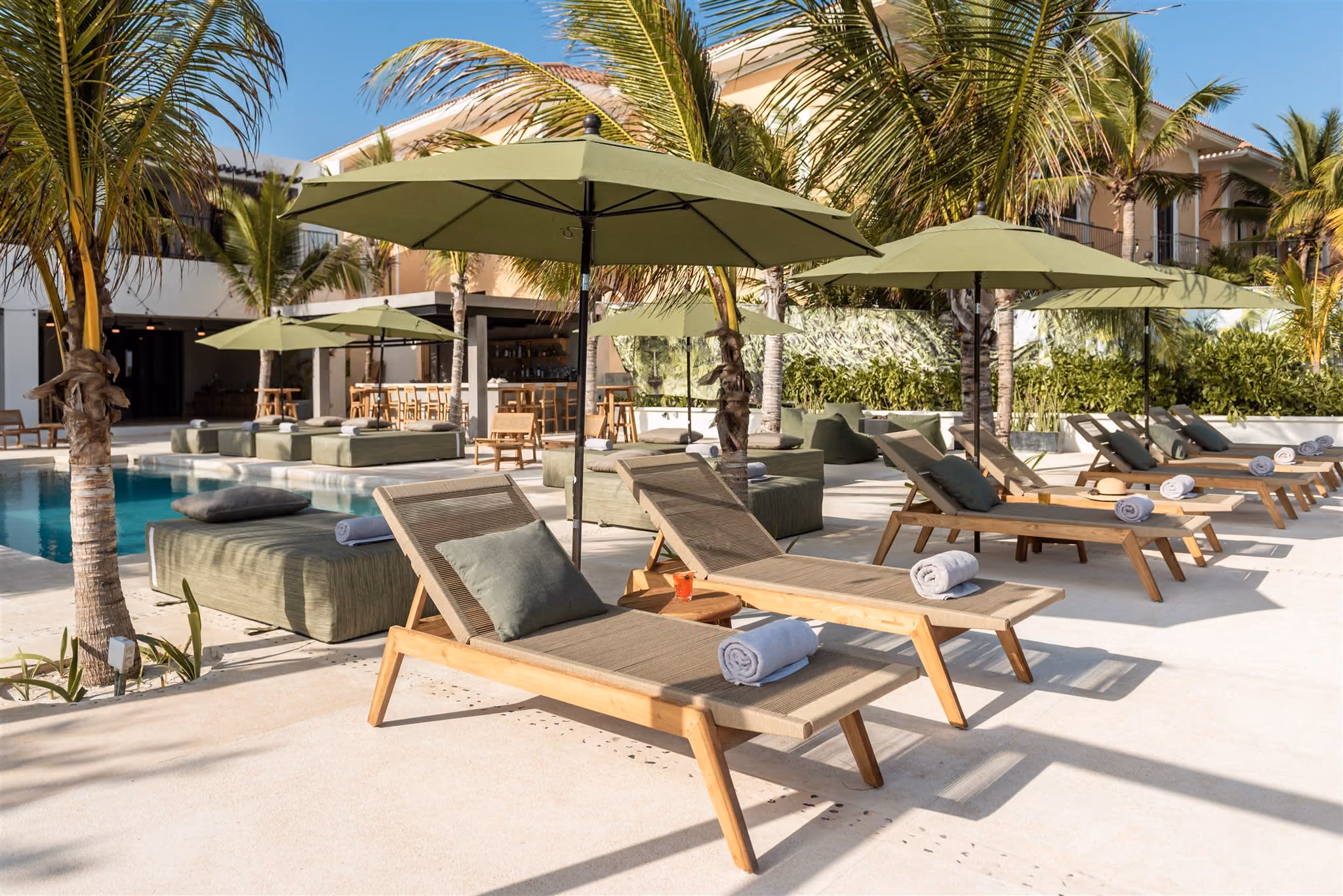 Outdoor pool with clear blue water, surrounded by palm trees, beige cushioned seating, and ocean view in the background.