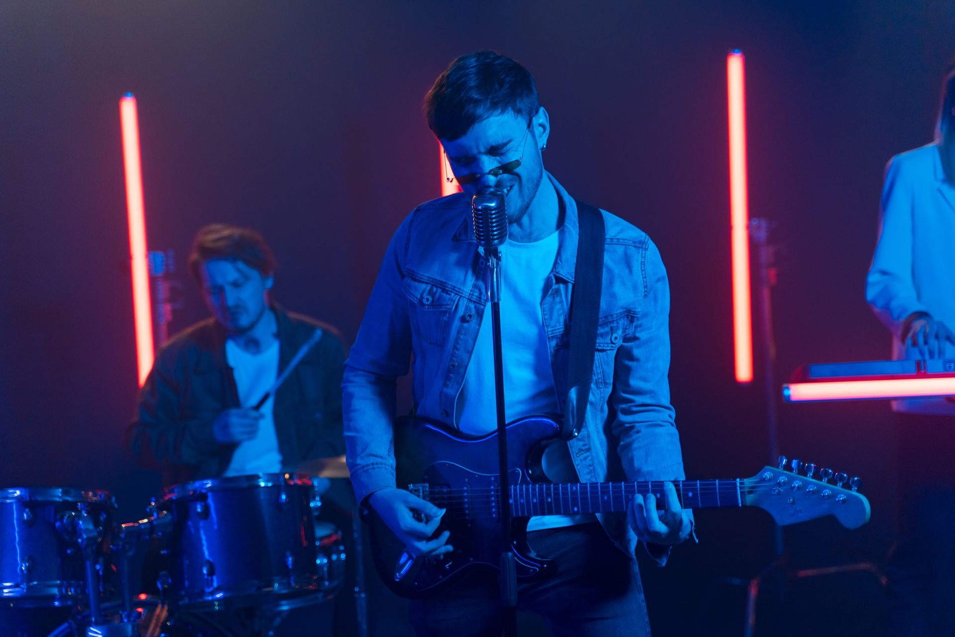Vibrant band performing with neon lights, featuring electric guitar and drums. Photo by AI25.Studio, via Pexels.com