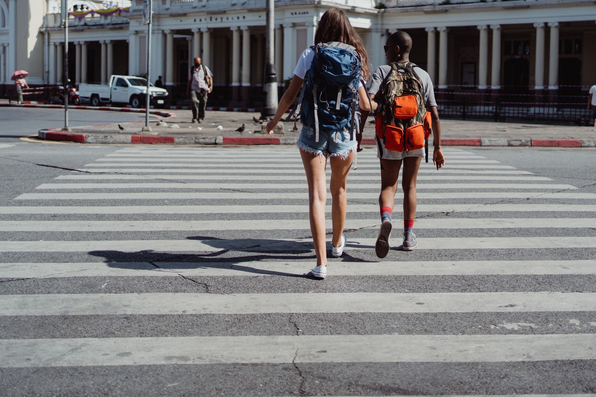 Two women with backpacks crossing a pedestrian lane in an urban setting on a sunny day. Photo by Ketut Subiyanto, via Pexels.com