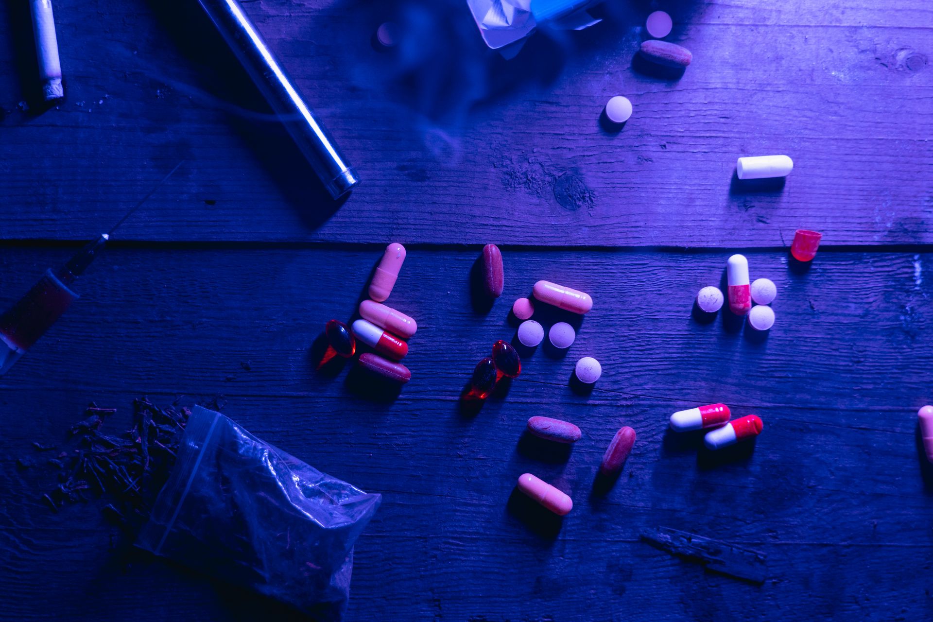 A flat lay of various drugs including pills, capsules, and a syringe on a wooden surface under moody blue lighting. Photo by MART PRODUCTION, via Pexels.com