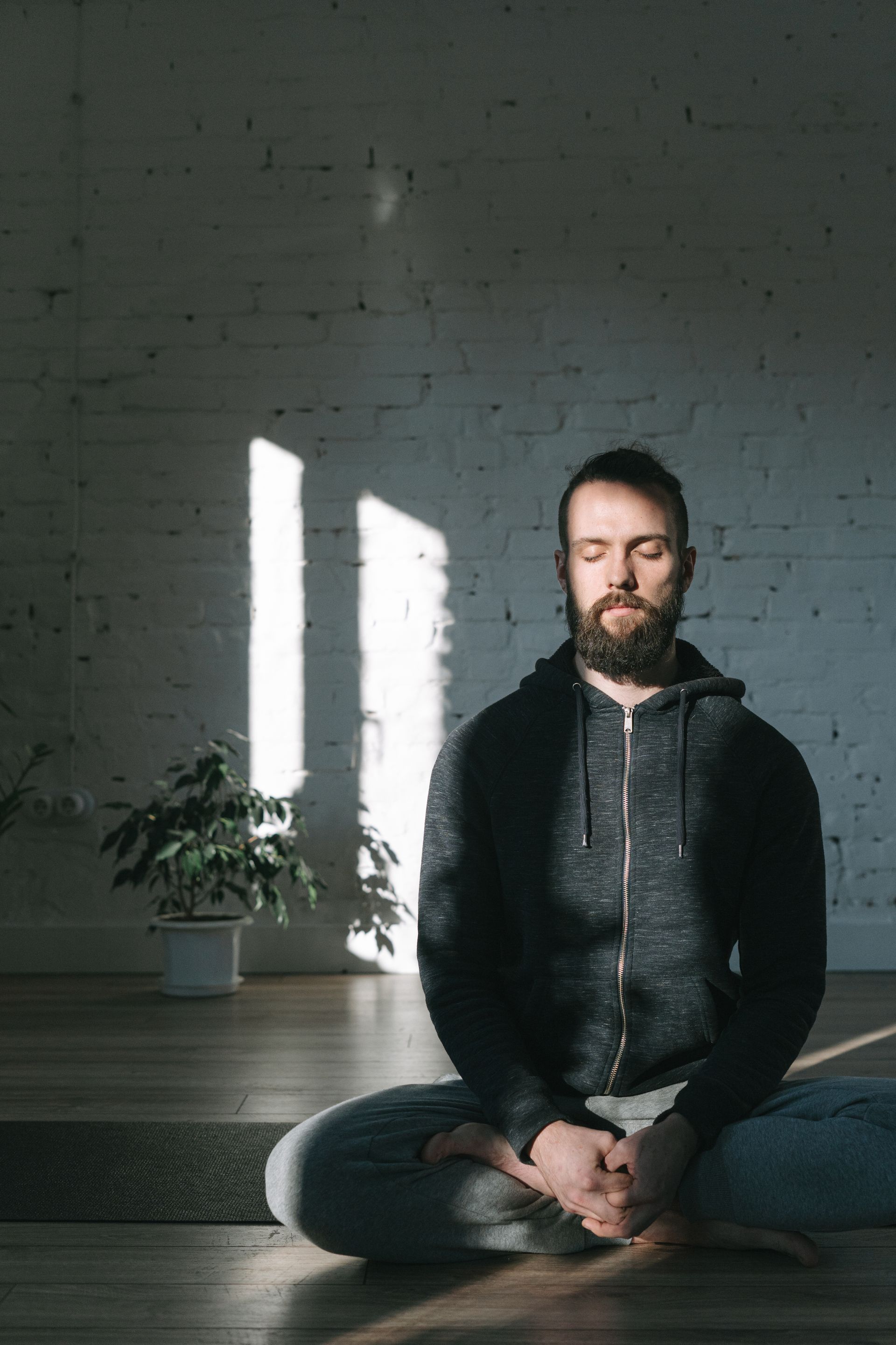 A man in a hoodie meditates in a sunlit yoga studio, embracing tranquility. Photo by Ivan S, via Pexels.com
