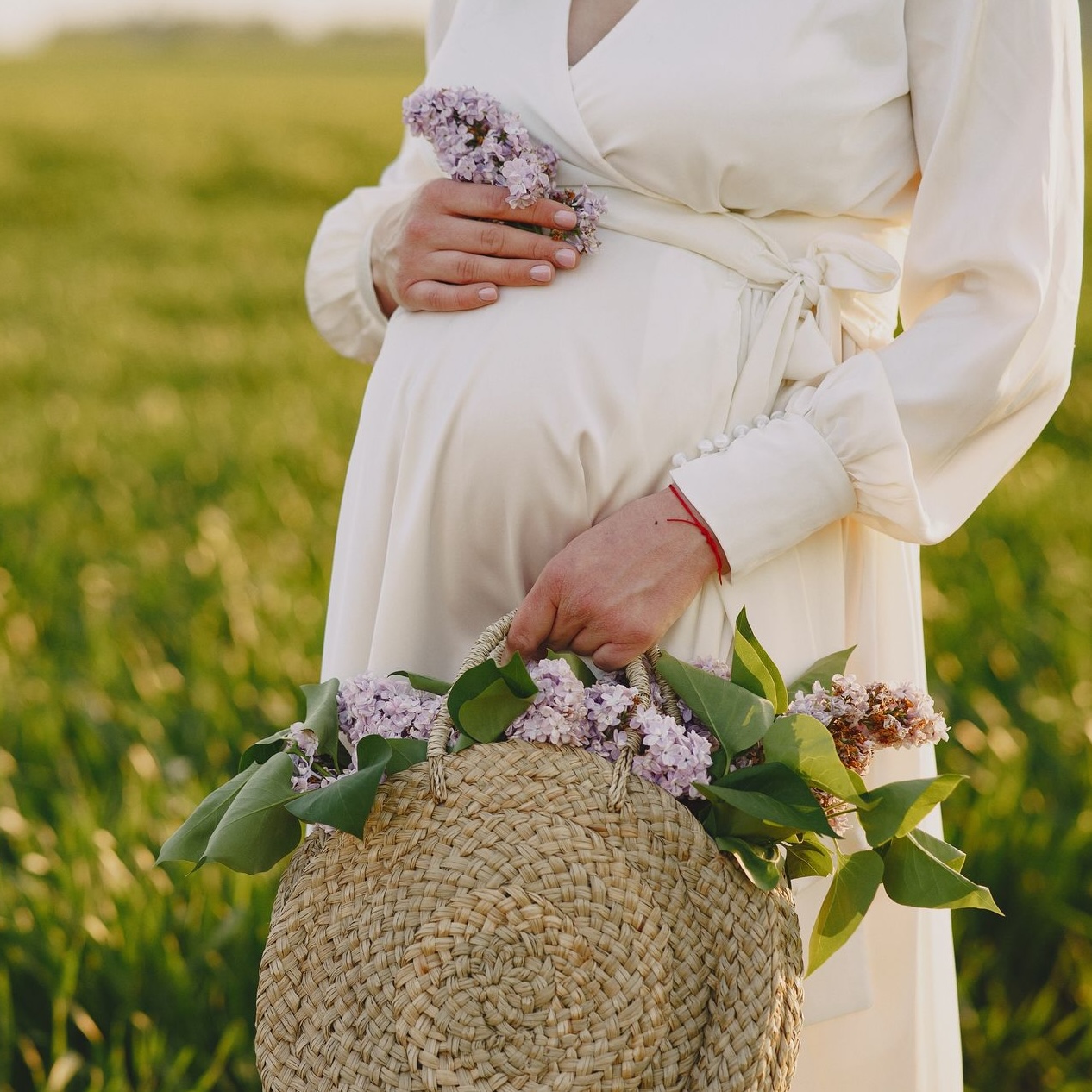 A pregnant woman in a white dress holds a straw bag with flowers in a lush green field. Photo by Gustavo Fring via Pexels.com