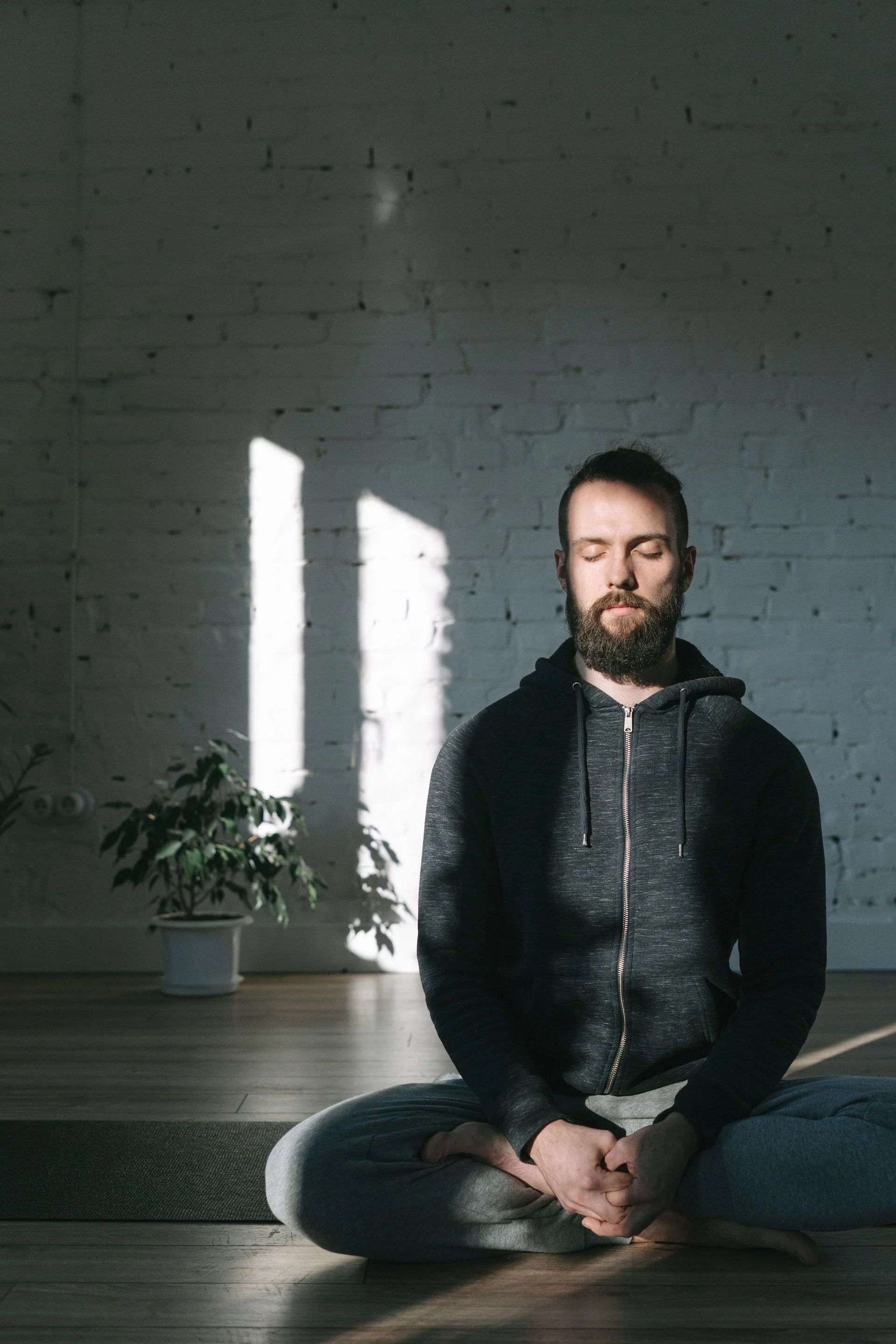 A man in a hoodie meditates in a sunlit yoga studio, embracing tranquility. Photo by Ivan S, via Pexels.com