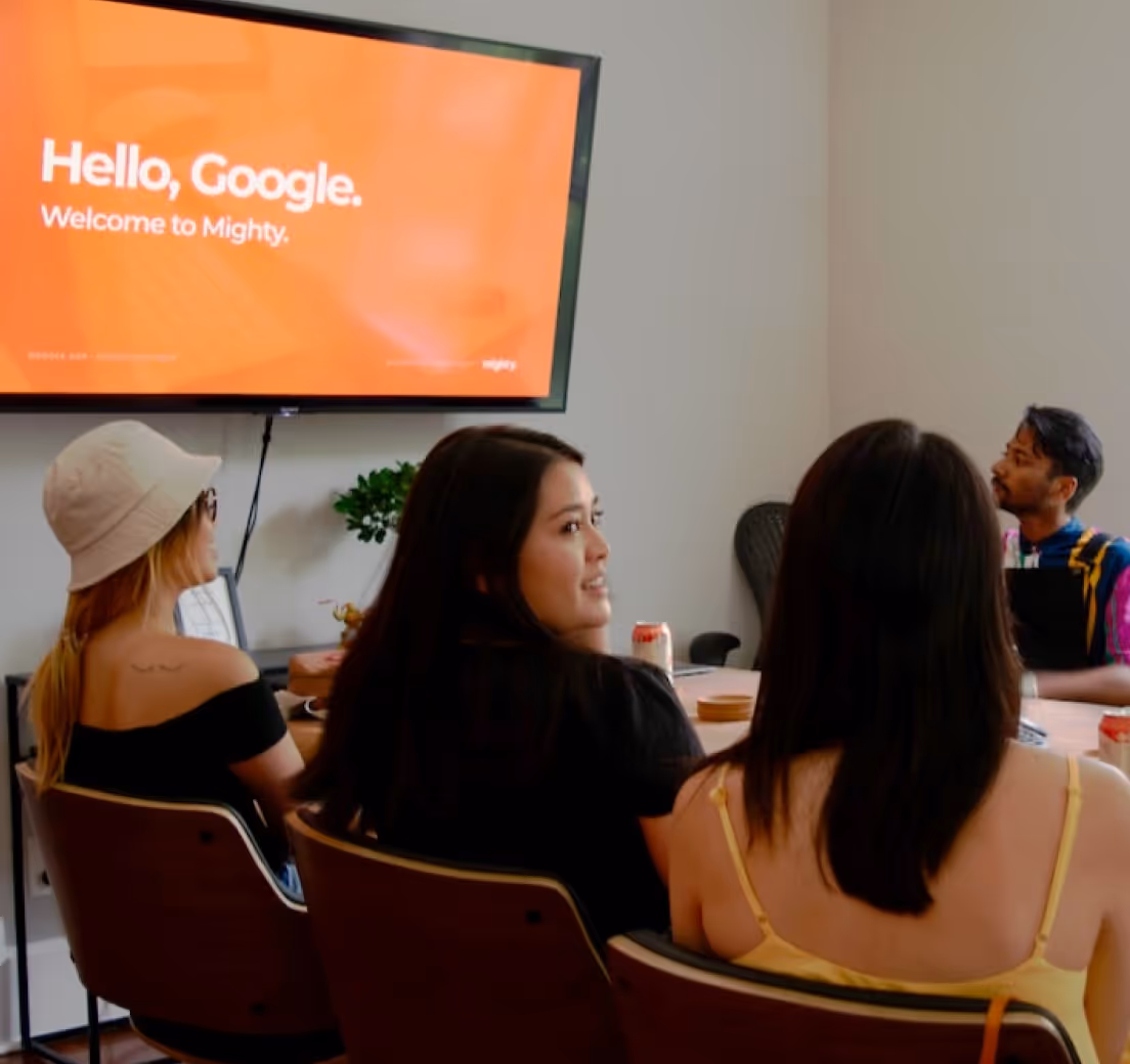Four people seated around a table watching a screen saying Hello, Google. Welcome to Mighty.