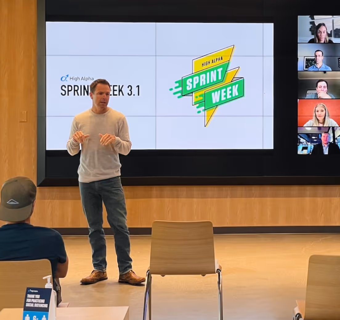 Man speaking to socially distanced attendees with Sprint Week presentation and remote participants on screen.
