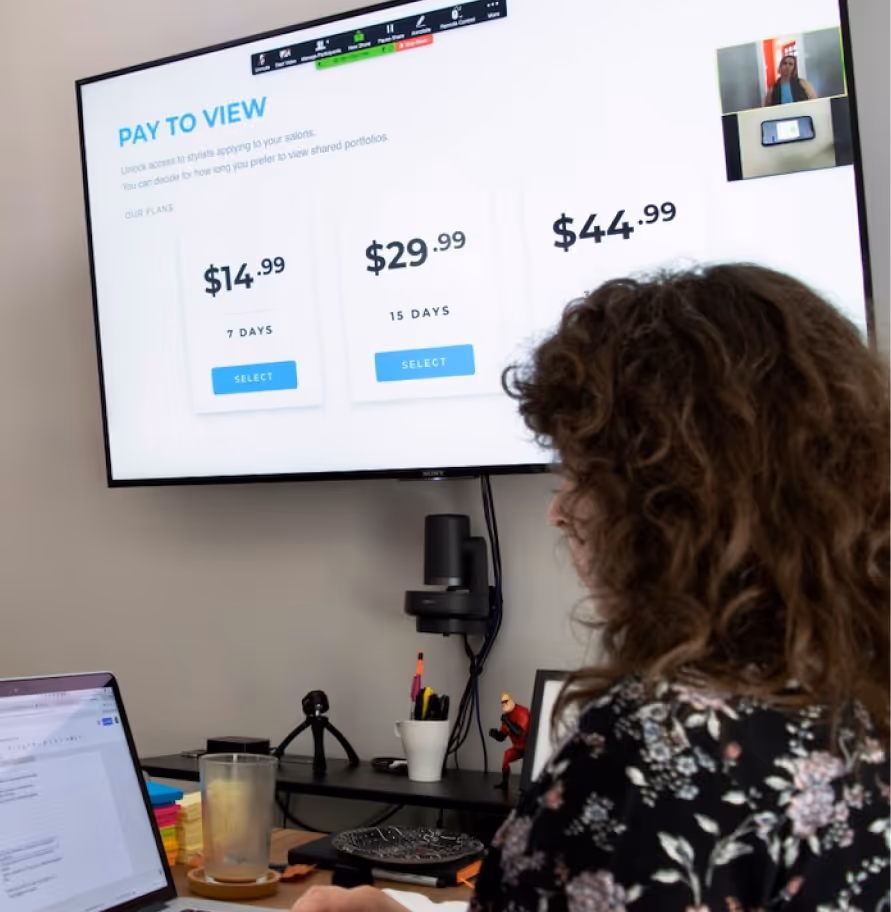 Person in floral shirt attending online meeting with pay-to-view pricing plans displayed on wall screen.