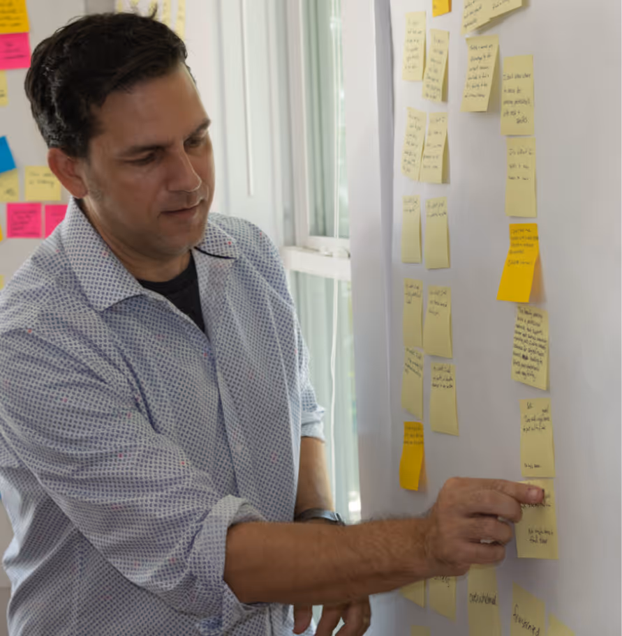 Man in patterned shirt pointing at sticky notes arranged on a whiteboard near a window.